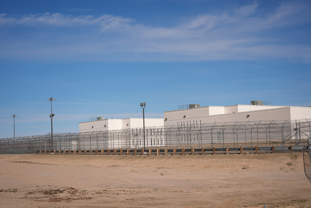 White prison buildings are surrounded by tall fences topped with barbed wire under a clear blue sky in a flat, sandy area.